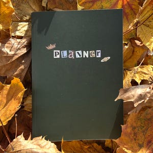 Könnte beinhalten: Ein dunkelgrüner Planer mit dem Wort "Planner" im Ausschnitt-Buchstabenstil, verziert mit einer kleinen Krone und einer Planetengrafik. Der Planer befindet sich vor einem Hintergrund aus Herbstblättern in Gelb- und Brauntönen.
