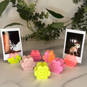 May include: A selection of flower-shaped soaps in various colours, including pink, orange, and yellow, each with a smiling face. Two polaroid-style photos of dogs are in the background. The soaps are displayed on a white marble surface.