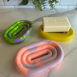May include: Three soap dishes in green, yellow, and marbled orange and purple. A bar of soap with the text "ALL-ONE!" sits on the yellow dish. The oval dishes have a grooved interior, offering a stylish way to store soap.