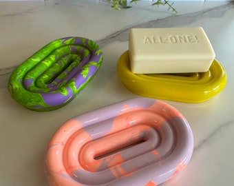 May include: Three oval soap dishes with a raised ribbed design. One dish features a green and purple marbled pattern, another is solid yellow with a rectangular off-white bar of soap embossed with the text ALL-ONE!, and the third is peach and lavender.