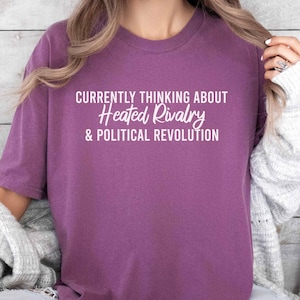 May include: A berry-colored short-sleeve t-shirt featuring the white printed text: CURRENTLY THINKING ABOUT Heated Rivalry & POLITICAL REVOLUTION. The shirt has a crew neckline and a relaxed fit, worn with a light gray cardigan and blue jeans.