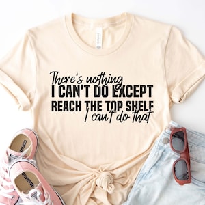 以下が含まれることがあります： 「There's nothing I CAN'T DO EXCEPT REACH THE TOP SHELF I can't do that」と黒文字で書かれたベージュのTシャツ。