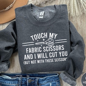 May include: Dark heather grey sweatshirt with white text that reads "Touch my fabric scissors and I will cut you (but not with these scissors)". The text is surrounded by a white line and a small scissor graphic.