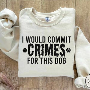 May include: Sand-colored sweatshirt with the text "I WOULD COMMIT CRIMES FOR THIS DOG" in black. The word "CRIMES" has a dripping effect, and paw prints flank the word. The sweatshirt is folded on a wooden surface.