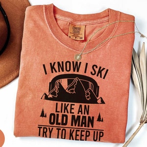 Könnte beinhalten: Ein korallenfarbenes T-Shirt mit dem Text "I KNOW I SKI LIKE AN OLD MAN TRY TO KEEP UP" in Schwarz. Das Shirt zeigt eine Berg-Grafik. Eine goldene Halskette und ein brauner Hut sind ebenfalls im Bild.