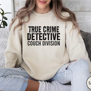 Pode incluir: Um moletom cor de areia com o texto preto "TRUE CRIME DETECTIVE COUCH DIVISION". O moletom é de cor neutra e tem um ajuste folgado. O texto está em uma fonte em negrito e desgastada.