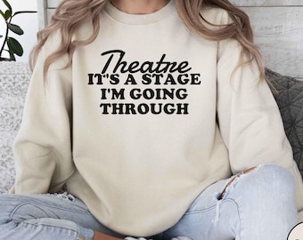 May include: A sand-coloured sweatshirt featuring the black text "Theatre IT'S A STAGE I'M GOING THROUGH". The sweatshirt has a crew neck and is a neutral colour. The word "SAND" is printed on a round label.