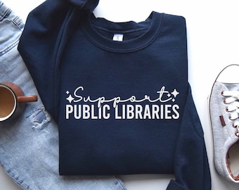 May include: A navy blue sweatshirt with the text "Support Public Libraries" in white lettering. The sweatshirt is laid flat on a white surface with a pair of gray sneakers and blue jeans.