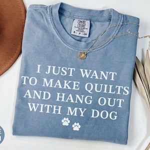 Puede incluir: Una camiseta de manga corta color azul jean con texto blanco que dice I JUST WANT TO MAKE QUILTS AND HANG OUT WITH MY DOG, acompañado de dos pequeños iconos de huellas de perro blancas debajo del texto. La camiseta tiene cuello redondo y etiqueta.