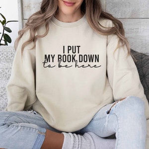 Op de afbeelding: Een beige sweatshirt met de tekst "I put my book down to be here" in het zwart gedrukt.