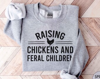 May include: A gray sweatshirt with the text "Raising Chickens and Feral Children" in black lettering. A black silhouette of a chicken is above the word "Chickens".