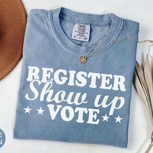 以下が含まれることがあります： REGISTER Show up VOTEという白いスクリーンプリントの文字と、VOTEという単語の両側に2つずつ星が描かれた、ブルーデニムカラーのTシャツ。シャツの内側の襟元にはComfort Colorsのブランドラベルがあり、クルーネック仕様になっています。