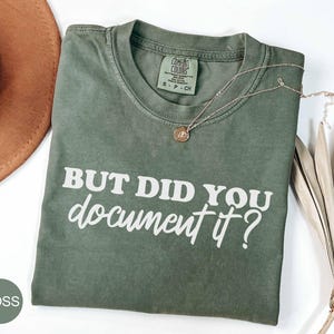 May include: A moss green t-shirt with the text "BUT DID YOU document it?" in white. A gold necklace with a pendant rests on the shirt. A brown hat and dried palm leaves are in the background.