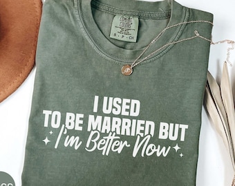 May include: A moss green t-shirt with the text "I USED TO BE MARRIED BUT I'm Better Now" in white. The shirt has a crew neck and a small tag on the upper left. A gold necklace with a pendant is draped across the shirt.