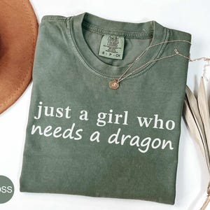 Puede incluir: Una camiseta de manga corta color verde musgo con cuello redondo y el texto blanco "just a girl who needs a dragon" impreso en la parte delantera. La camiseta tiene una etiqueta de la marca Comfort Colors en el cuello interior y es talla S.