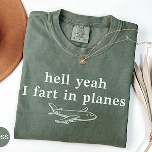 May include: A moss green short-sleeved t-shirt featuring the white text "hell yeah I fart in planes" and a line drawing of an aeroplane. The shirt has a Comfort Colors brand label at the inner neckline and is made of cotton.
