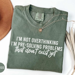 May include: A moss green short-sleeve t-shirt featuring the white printed text: I'M NOT OVERTHINKING I'M PRE-SOLVING PROBLEMS that don't exist yet. The shirt has a crew neckline and a Comfort Colors brand label on the inside collar area.
