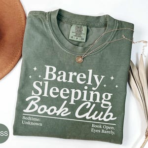 Könnte beinhalten: Ein moosgrünes Kurzarm-T-Shirt mit weißem Text, der Barely Sleeping Book Club, Bedtime: Unknown und Book Open. Eyes Barely lautet. Das T-Shirt hat ein Comfort Colors-Etikett und ist mit kleinen weißen Sternsymbolen verziert.