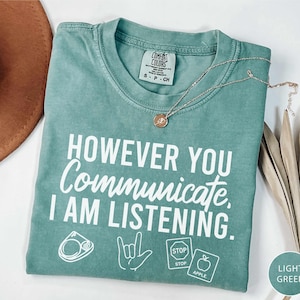 May include: Light green t-shirt with the text "HOWEVER YOU Communicate, I AM LISTENING." and graphic icons. A gold necklace and a brown hat are also visible. The shirt is made of a soft material.