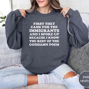 May include: Dark heather gray sweatshirt with the text "FIRST THEY CAME FOR THE IMMIGRANTS AND I SPOKE UP BECAUSE I KNOW THE REST OF THE GODDAMN POEM" in white block letters. The sweatshirt is a casual style.