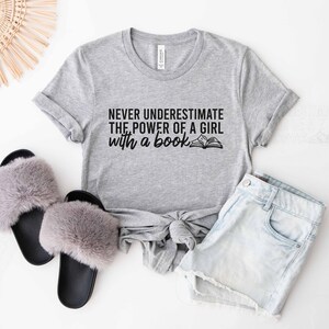 An athletic heather color shirt with the saying "Never underestimate the power of a girl with a book"