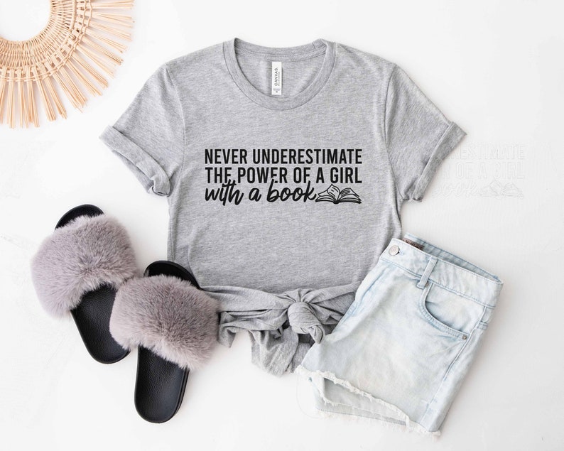An athletic heather color shirt with the saying "Never underestimate the power of a girl with a book"