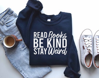 May include: A navy blue sweatshirt with the text "READ BOOKS BE KIND STAY WEIRD" in white lettering. The sweatshirt is laid flat on a white surface with a pair of light gray sneakers and a pair of blue jeans.