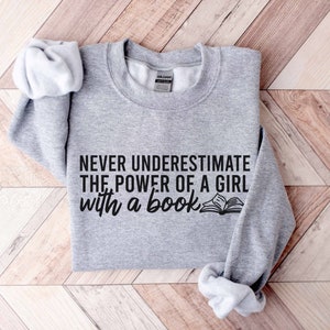 A sport grey color sweatshirt with the saying "Never underestimate the power of a girl with a book"