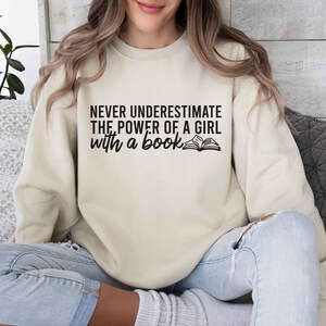 A sand color sweatshirt with the saying "Never underestimate the power of a girl with a book"