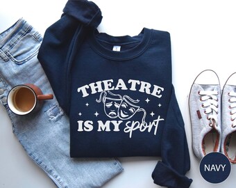 May include: A navy blue sweatshirt featuring the white text "THEATRE IS MY sport" and comedy/tragedy masks. The image includes light blue jeans, a coffee cup, and grey trainers. The sweatshirt is a casual item.