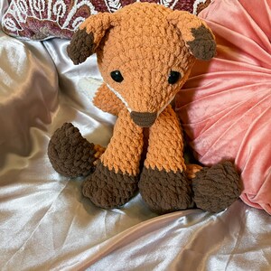 May include: A handmade orange and brown crochet fox stuffed animal. The fox has dark brown paws, ears, and nose, with black safety eyes. The plush toy is sitting on a satin surface.