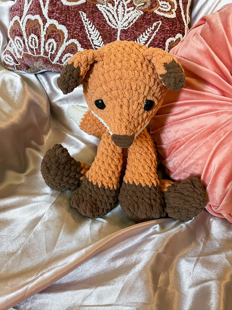 May include: A handmade orange and brown crochet fox stuffed animal. The fox has dark brown paws, ears, and nose, with black safety eyes. The plush toy is sitting on a satin surface.