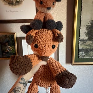 May include: Two handmade fox plush toys. The larger fox is held up, showcasing its orange and brown crochet body. The smaller fox sits on top, with similar coloring. The toys have black accents on their ears and paws, and white accents on their faces.