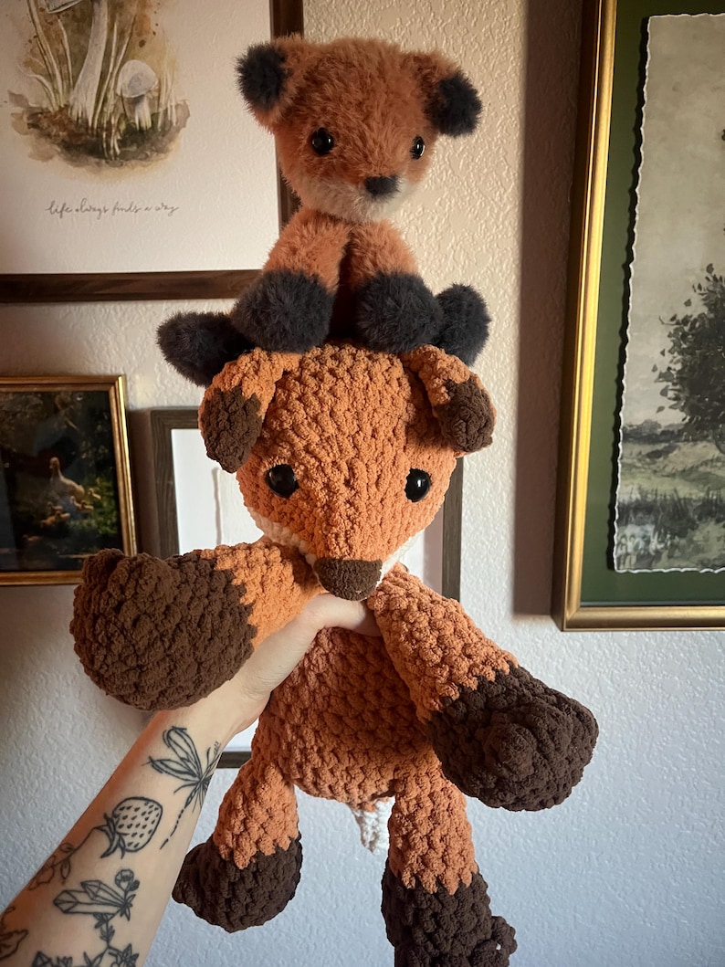 May include: Two handmade fox plush toys. The larger fox is held up, showcasing its orange and brown crochet body. The smaller fox sits on top, with similar coloring. The toys have black accents on their ears and paws, and white accents on their faces.