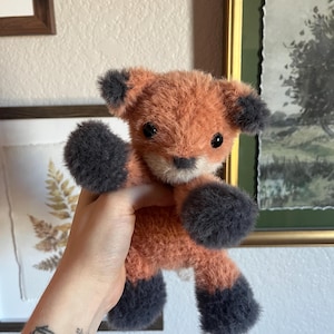 May include: A handmade plush fox toy with orange and gray fur. The fox has dark gray paws, ears, and a small nose. The toy is held up in front of a neutral background, showcasing its soft, textured appearance. The fox is a cute, cuddly toy.
