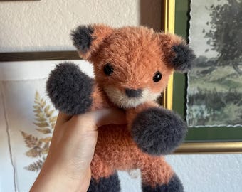 May include: A handmade plush fox toy with orange and gray fur. The fox has dark gray paws, ears, and a small nose. The toy is held up in front of a neutral background, showcasing its soft, textured appearance. The fox is a cute, cuddly toy.