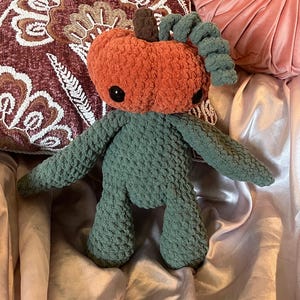 May include: A handmade crochet pumpkin doll with a plush orange head, dark green body, and black button eyes. The doll has a brown stem and green curly vines. The doll is sitting on a pink satin surface.