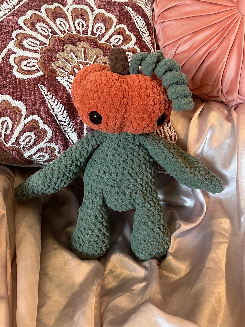 May include: A handmade crochet pumpkin doll with a plush orange head, dark green body, and black button eyes. The doll has a brown stem and green curly vines. The doll is sitting on a pink satin surface.