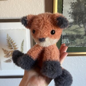 May include: A handmade plush fox toy with orange and gray fur. The fox has black eyes, a white muzzle, and gray paws and ears. The toy is held in a hand, showcasing its soft texture and detailed craftsmanship.
