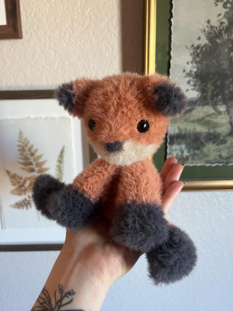 May include: A handmade plush fox toy with orange and gray fur. The fox has black eyes, a white muzzle, and gray paws and ears. The toy is held in a hand, showcasing its soft texture and detailed craftsmanship.