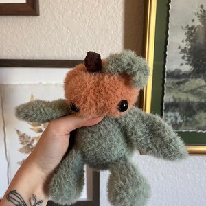 May include: A handmade plush toy in the shape of a bear. The bear has a peach-colored head with a small brown top and green fur for the body and limbs. The bear has black button eyes.