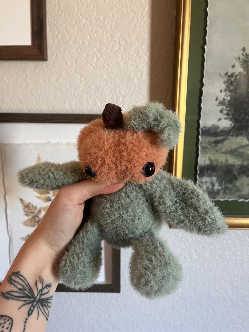 May include: A handmade plush toy in the shape of a bear. The bear has a peach-colored head with a small brown top and green fur for the body and limbs. The bear has black button eyes.