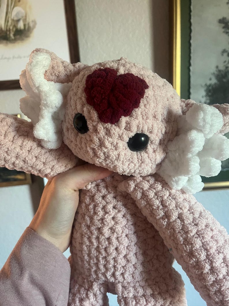 May include: A handmade, light pink crochet stuffed animal with a burgundy heart on its head. The toy has large black eyes and white fluffy accents. The plushie is soft and cuddly, perfect for a child's bedroom or nursery.