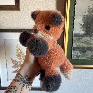 May include: A plush fox toy with orange fur, gray paws, and ears. The fox has a white chest and a black button eye. The toy is held up against a wall with framed artwork.