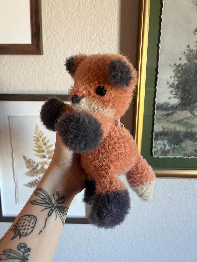 May include: A plush fox toy with orange fur, gray paws, and ears. The fox has a white chest and a black button eye. The toy is held up against a wall with framed artwork.