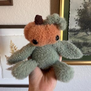 May include: A handmade plush toy in the shape of a pumpkin. The pumpkin has an orange face and a brown stem. The body and limbs are a soft green color. The toy has black button eyes and is held in a hand.