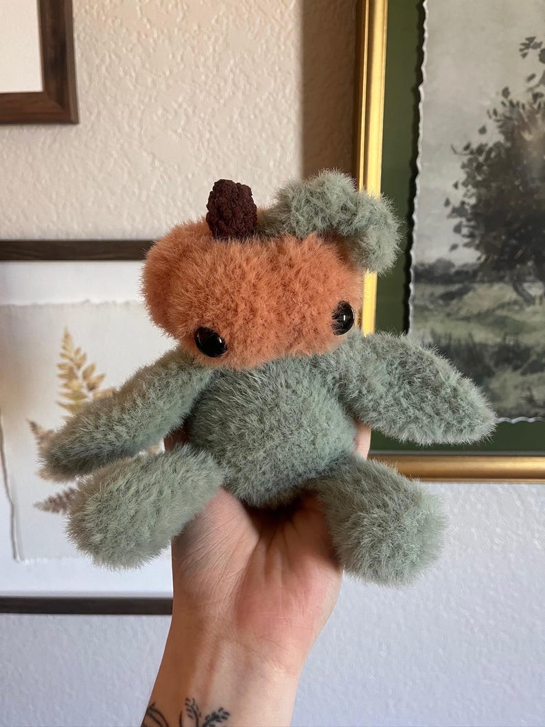 May include: A handmade plush toy in the shape of a pumpkin. The pumpkin has an orange face and a brown stem. The body and limbs are a soft green color. The toy has black button eyes and is held in a hand.