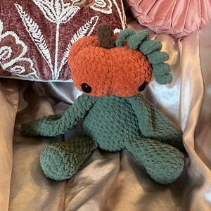 May include: A handmade crochet pumpkin plush toy with an orange body, green stem and leaves, and black eyes. The plush has green arms and legs. The toy is sitting on a satin surface with decorative pillows in the background.