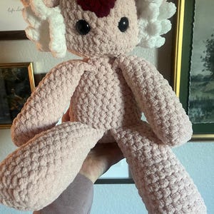May include: A handmade, light pink crochet axolotl plush toy. It features large black eyes, white frills, and a dark red heart-shaped detail on its head. The plush has a soft, textured appearance.