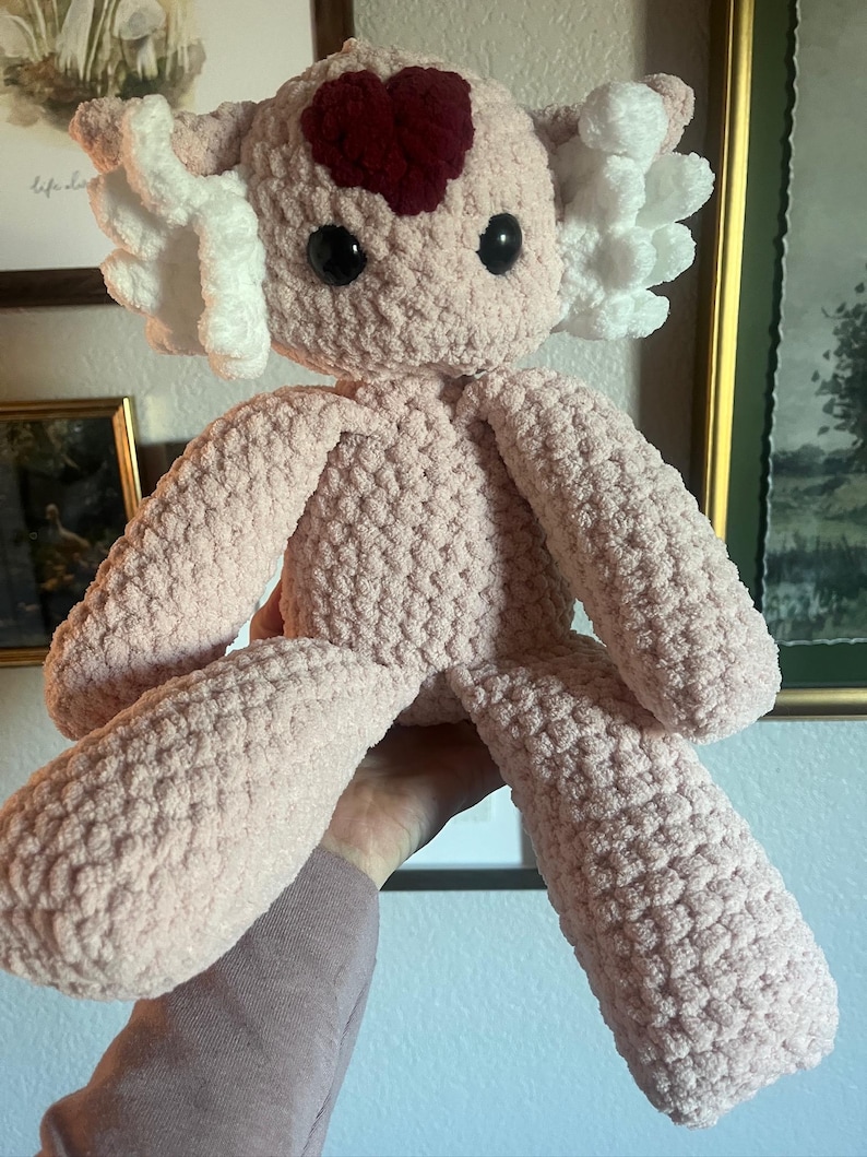 May include: A handmade, light pink crochet axolotl plush toy. It features large black eyes, white frills, and a dark red heart-shaped detail on its head. The plush has a soft, textured appearance.